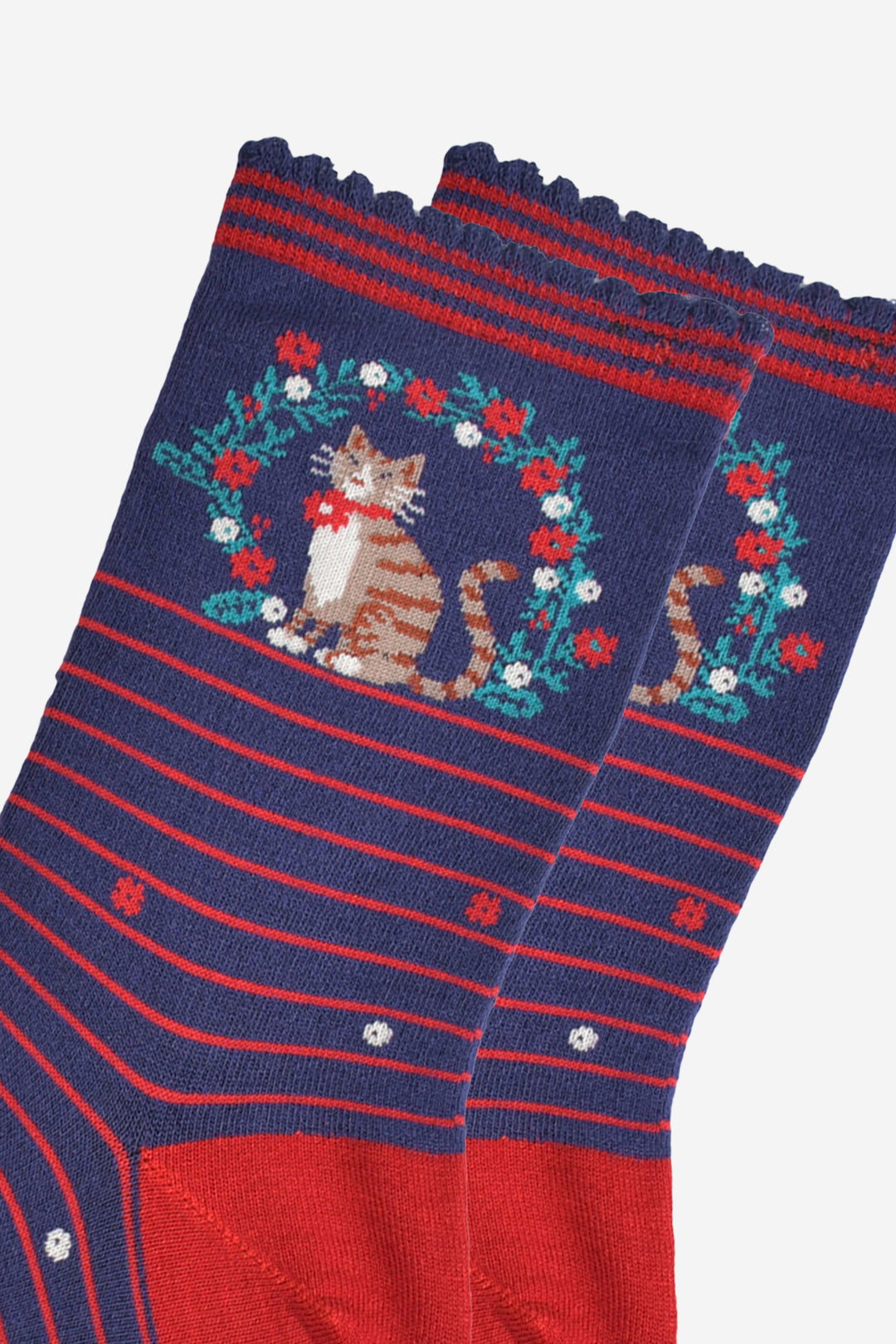 close up of the cat and wreath, the cat is tabby, light brown with stripes, it has a white belly and face and is wearing a festive red bow on its collar. the wreath is green with white and red flowers.