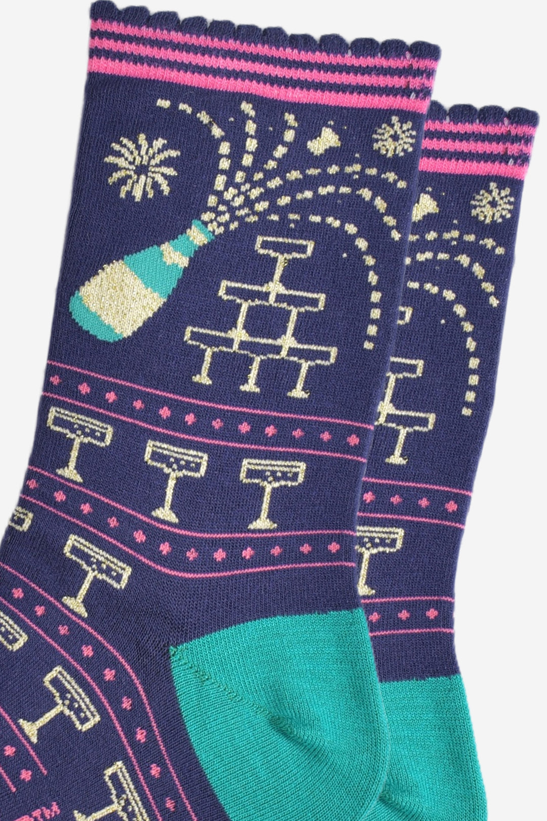 close up of the champagne design on the ankle of the socks, there is a green and gold champagne bottle with a cork flying out of it and spraying the sparkling wine all over. there is a pyramid of champagne coupe glasses and gold fireworks in the background