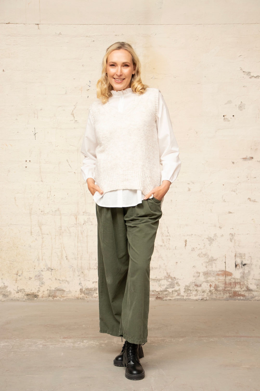 model wearing green khaki cord trousers with pocket, paired with black ankle boots and a relaxed fitting cream knitted vest and white shirt.