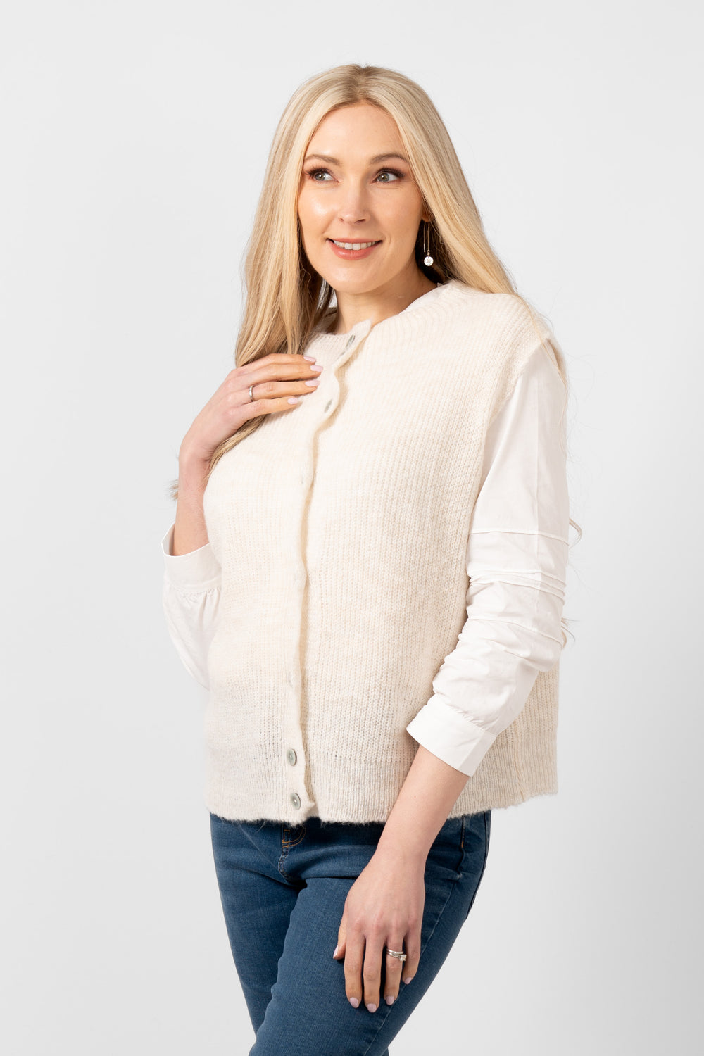 sleeveless cream knitted tank top shown styled over a white blouse and paired with blue jeans.
