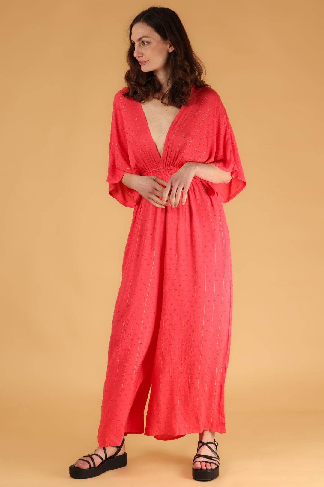 model wearing a coral wide leg jumpsuit with a deep v neck and a gold metallic stripe 