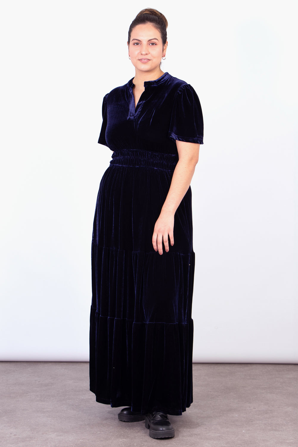 model wearing a maxi tiered velvet dress in navy blue, the dress has short sleeves and an open v-neck collar