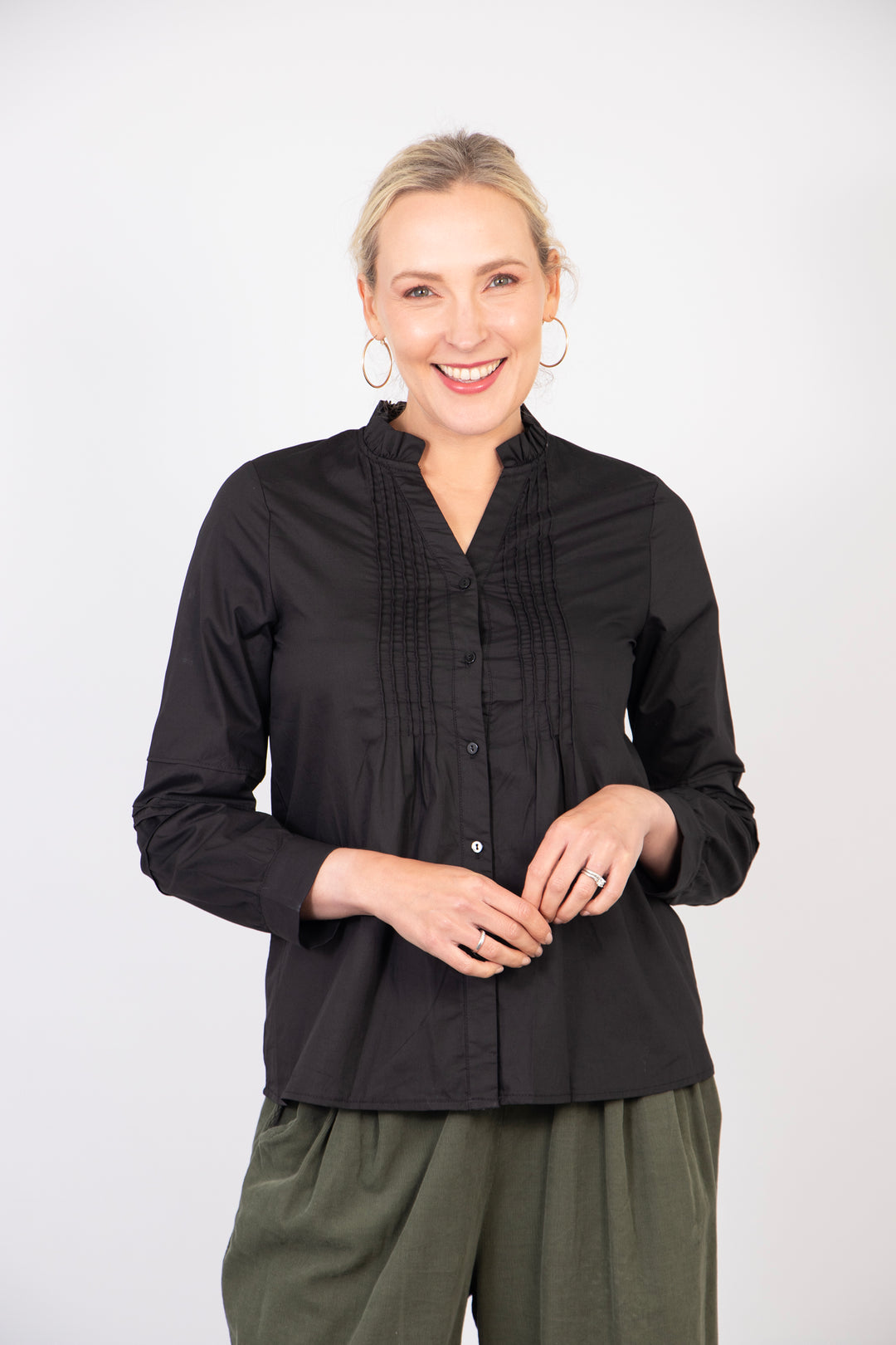 model wearing a black cotton long sleeve blouse. the blouse has small pleats on the front on each side of the neckline. the collar is open and has a ruffle trim