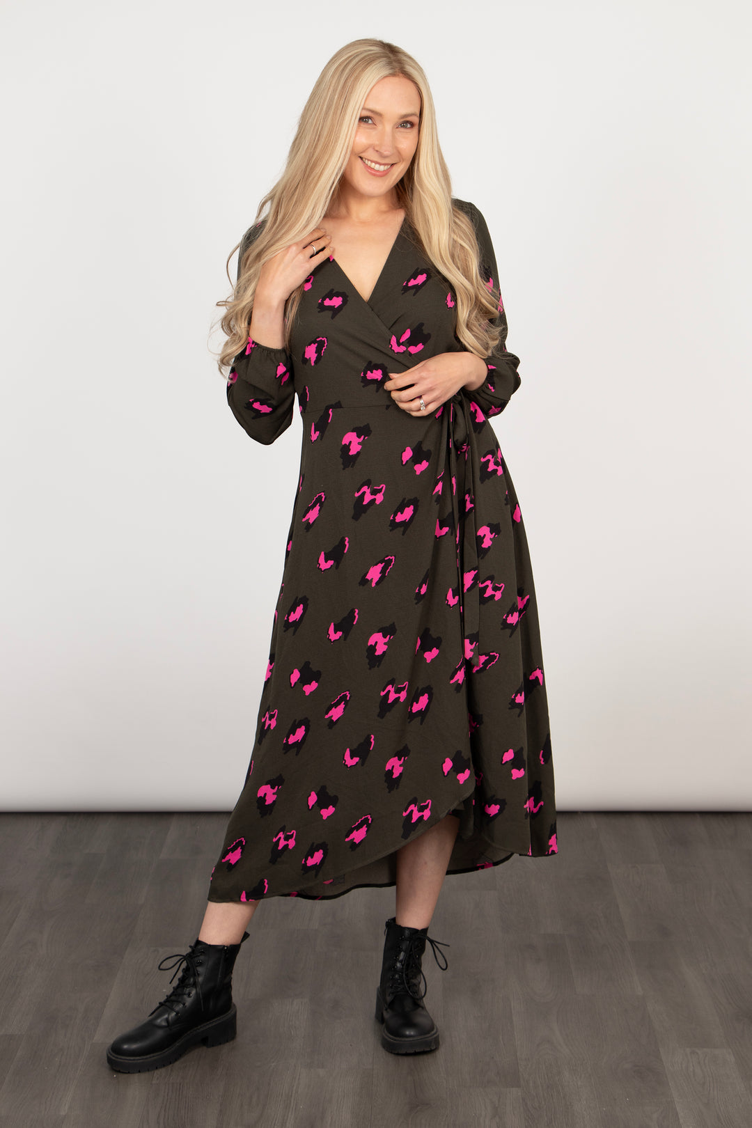 model wearing a khaki and pink dipped hem wrap dress paired with black ankle boots. the dress has a v-neck, waist tie and long sleeves