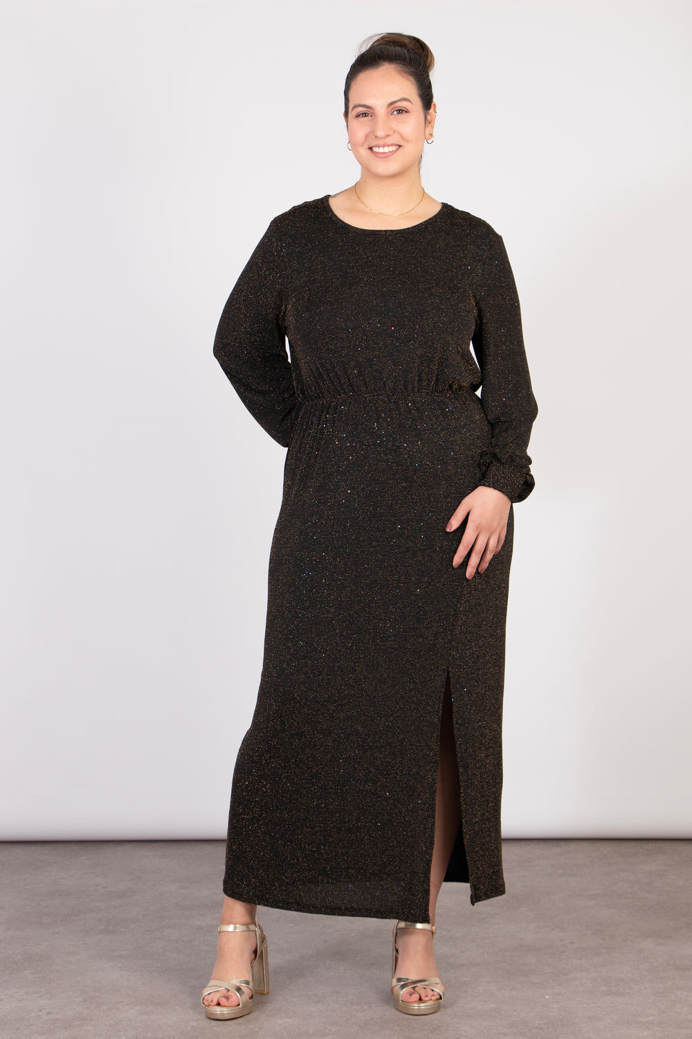 model wearing a black and bronze sparkly glitter dress with a knee high split at the front and a cinched elasticated waist. the dress has long sleeves and the glitter sparkle is all over the design