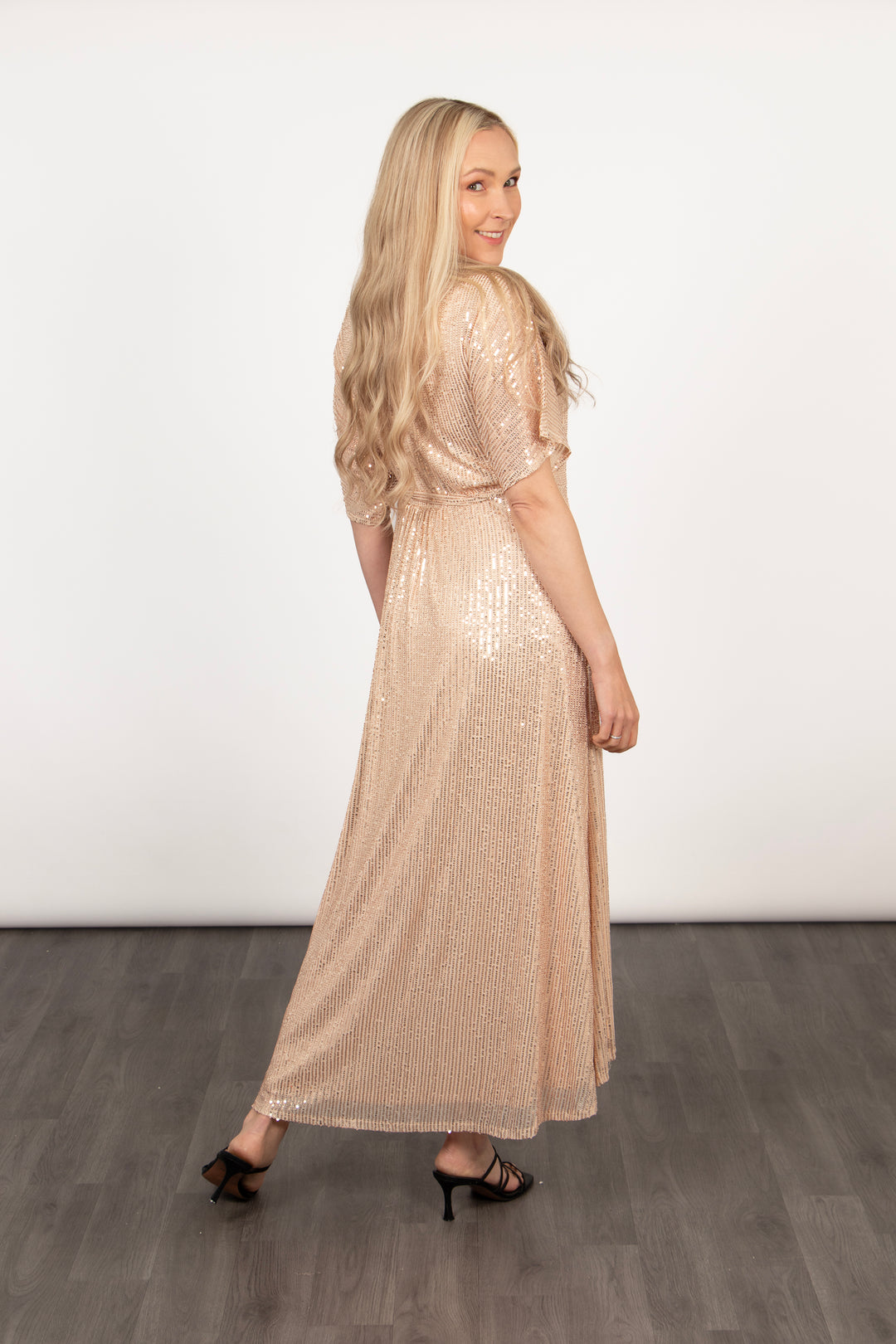 model showing the back of the gold sequin wrap dress, showing that the back of the dress is longer than the front. the back is maxi length and sits at the ankle