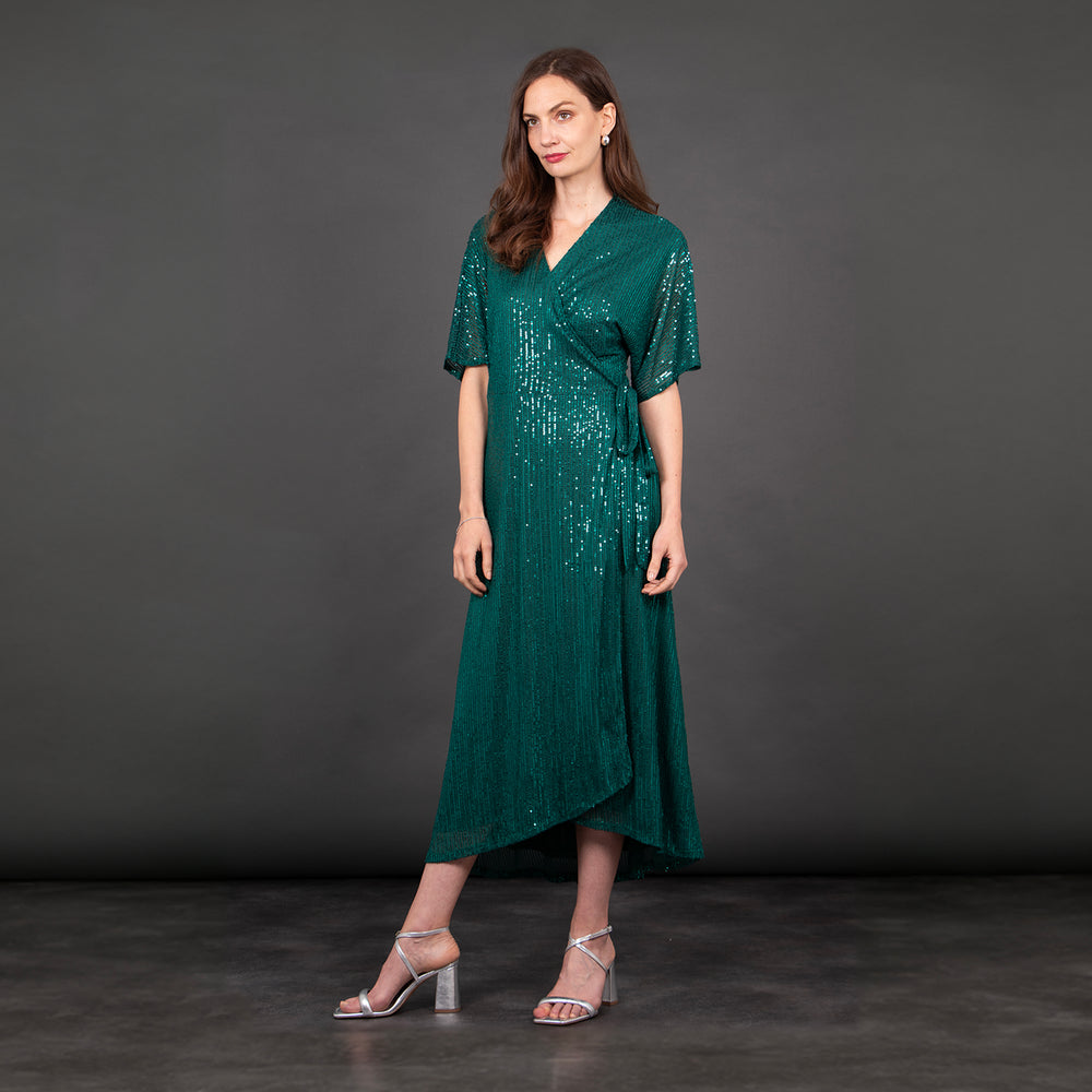 model wearing a dipped hem wrap dress in green with an all over sequin sparkle. the dress has a v-neck and waist tie