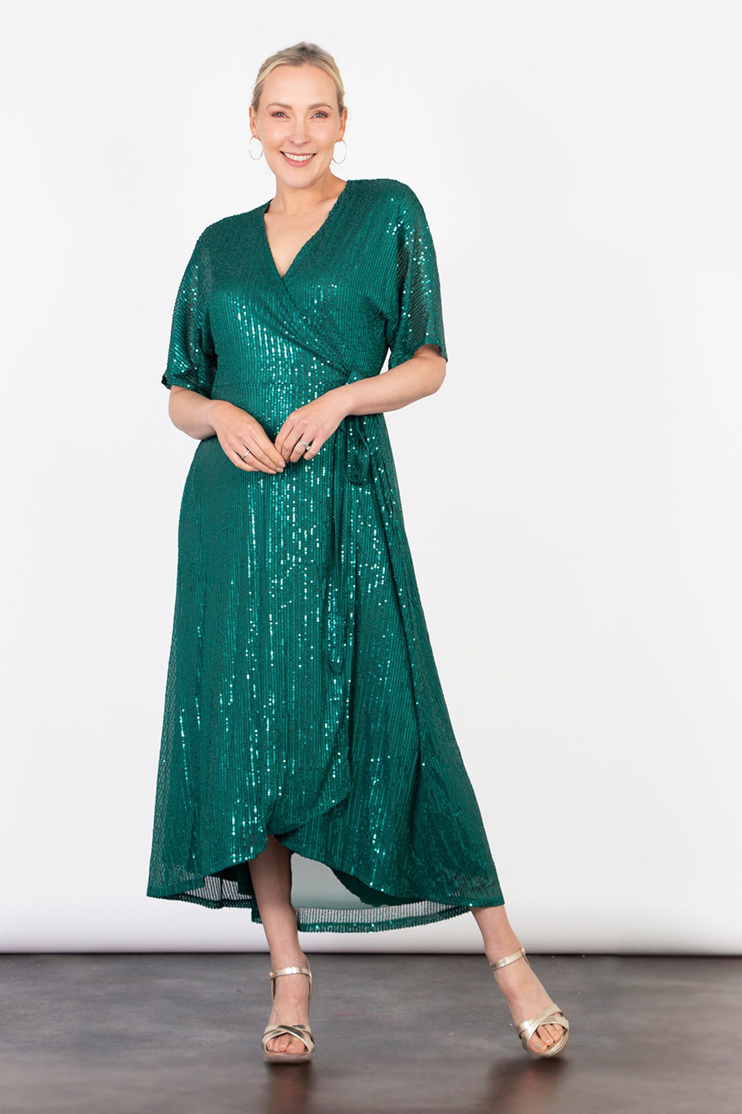 model wearing a green midi length dipped hem sparkly sequinned wrap dress with a v-neck and waist tie fastening