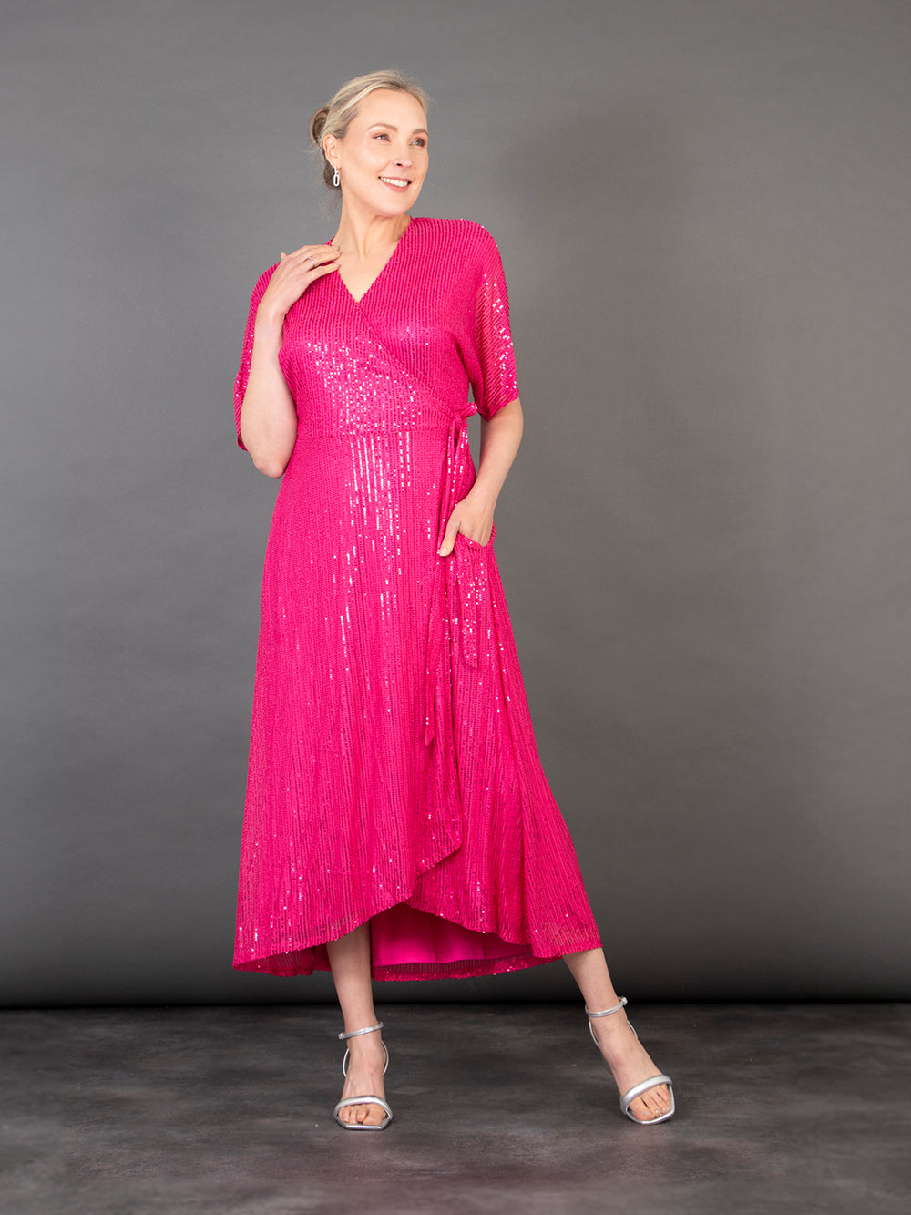 model wearing a pink midi length dipped hem sparkly sequinned wrap dress with a v-neck and waist tie fastening