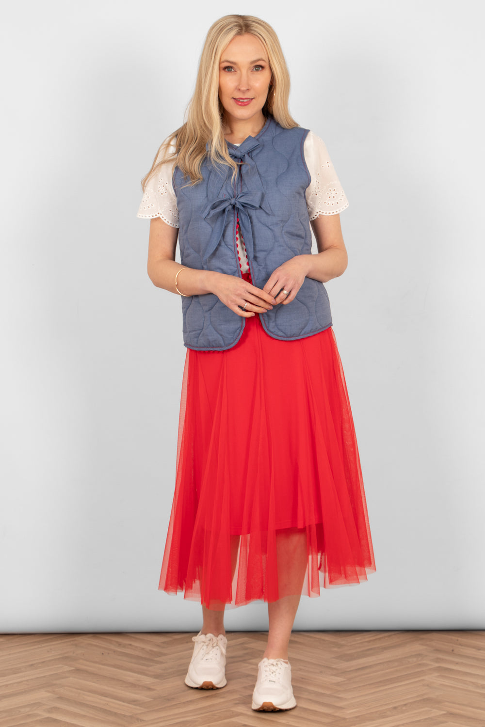 model wearing a red tulle skirt styled with a blue sleeveless gilet, white short sleeve blouse and white trainers showing a casual look