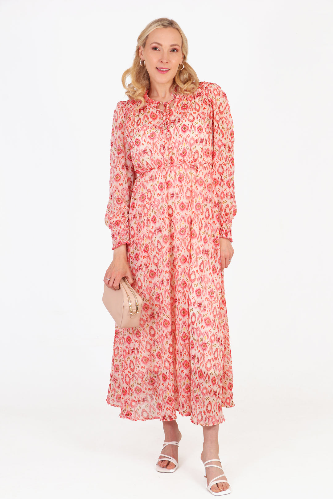 model wearing coral pink aztec pattern long sleeve dress, paired with sandals and a beige leather bag. the dress has a ruffled neck tie collar and an all over pattern