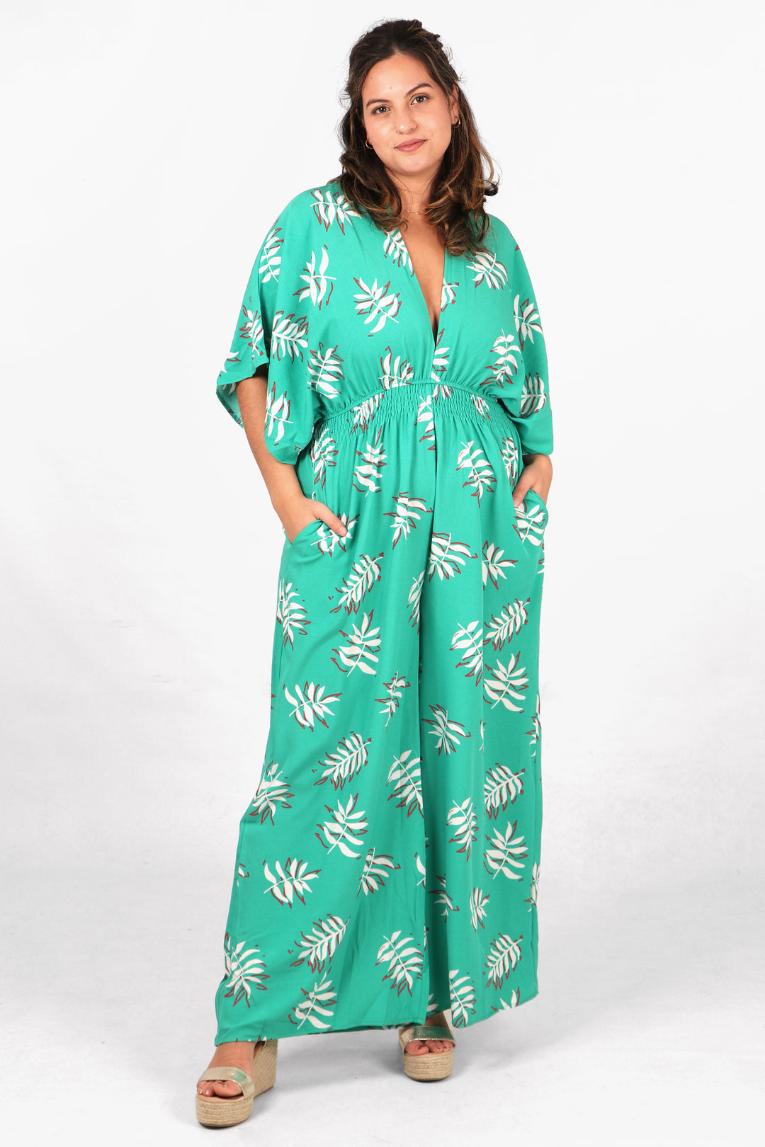 model wearing a mint green jumpsuit with an all over pattern of white palm leaves. the jumpsuit has a deep v neck, elbow length angel sleeves and a shirred waist