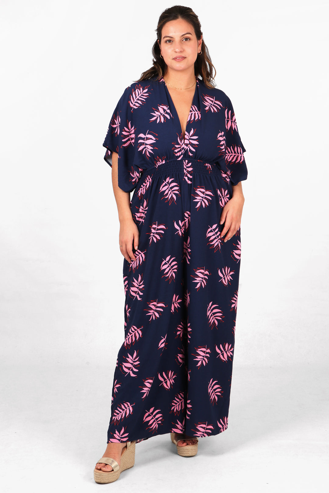 model wearing a navy blue jumpsuit with an all over pattern of pink palm leaves. the jumpsuit has a deep v neck, elbow length angel sleeves and a shirred waist