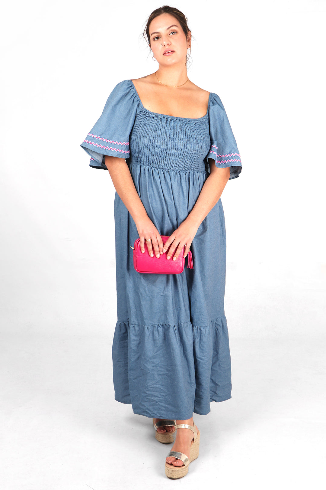 model wearing a denim blue maxi milkmaid dress with shirred bodice and bell sleeves, the sleeves have pink zig zag embroidered stripes around the cuffs
