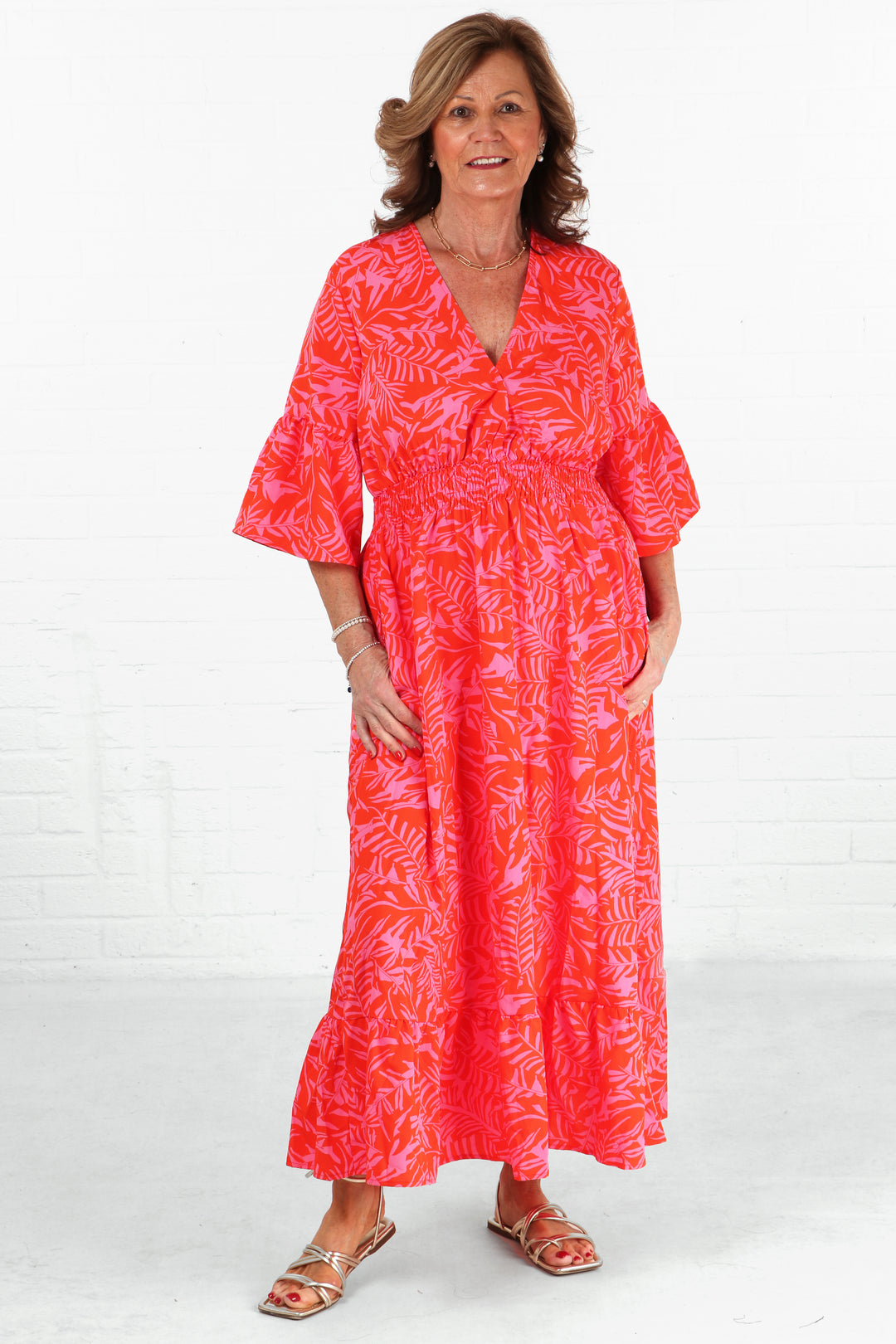 model wearing a maxi length v neck dress with loose fluted sleeves, vneck and a shirrred waist in an all over pink and orange tropical leaf print pattern