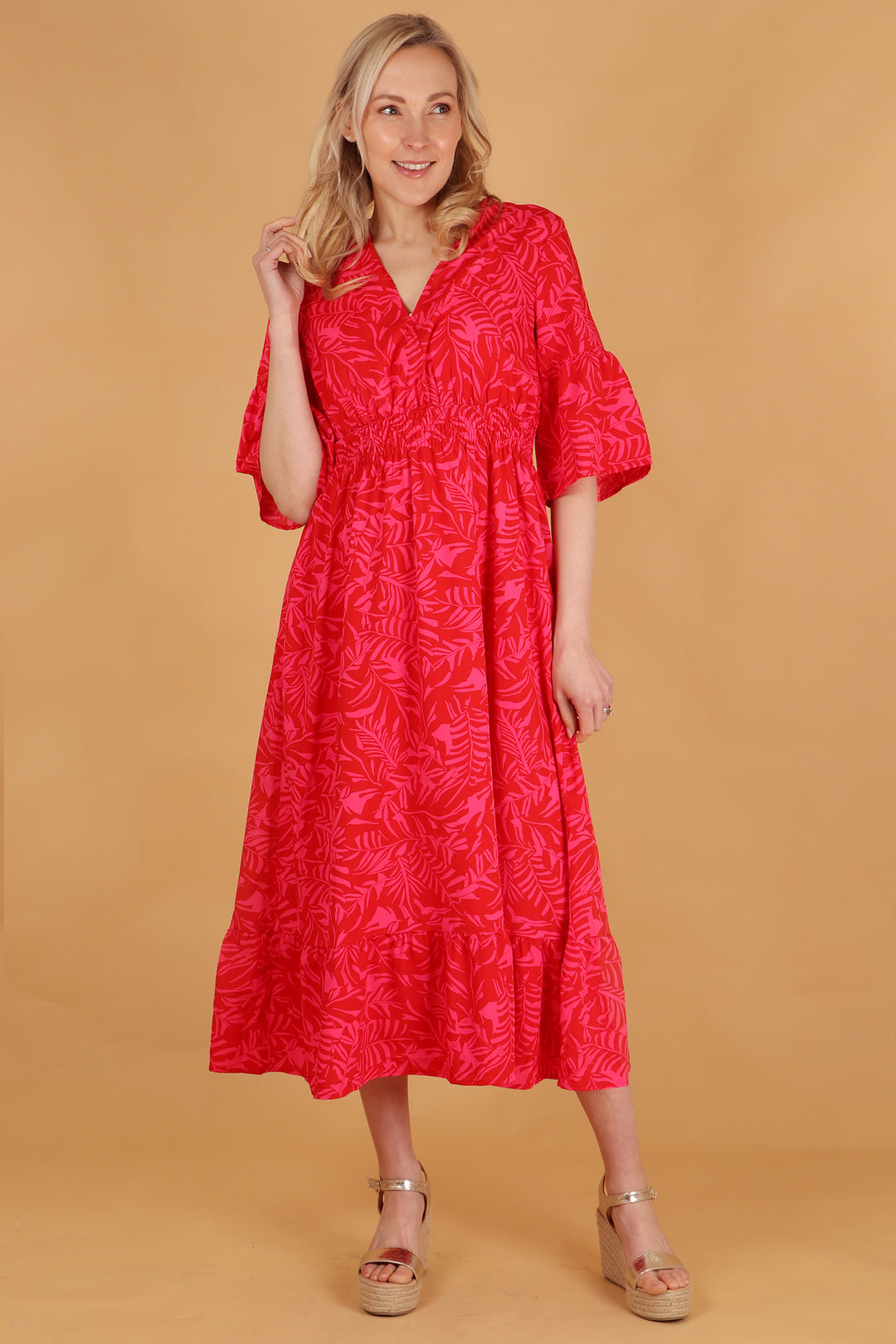 model wearing a midaxi length red and pink tropical leaf pattern v neck dress with a shirred waist and 3/4 sleeves