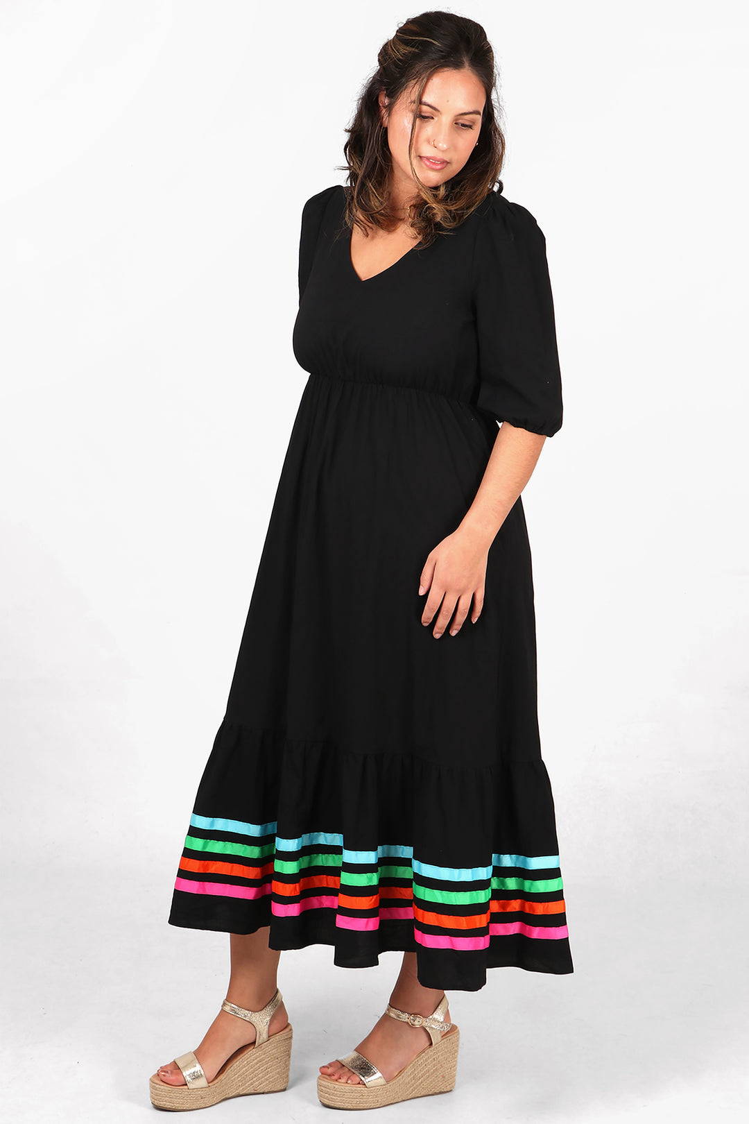 model wearing a black midaxi length dress with four horizontal rainbow ribbon stripes on the bottom tier of the dress, dress has a v neck and elbow length short sleeves
