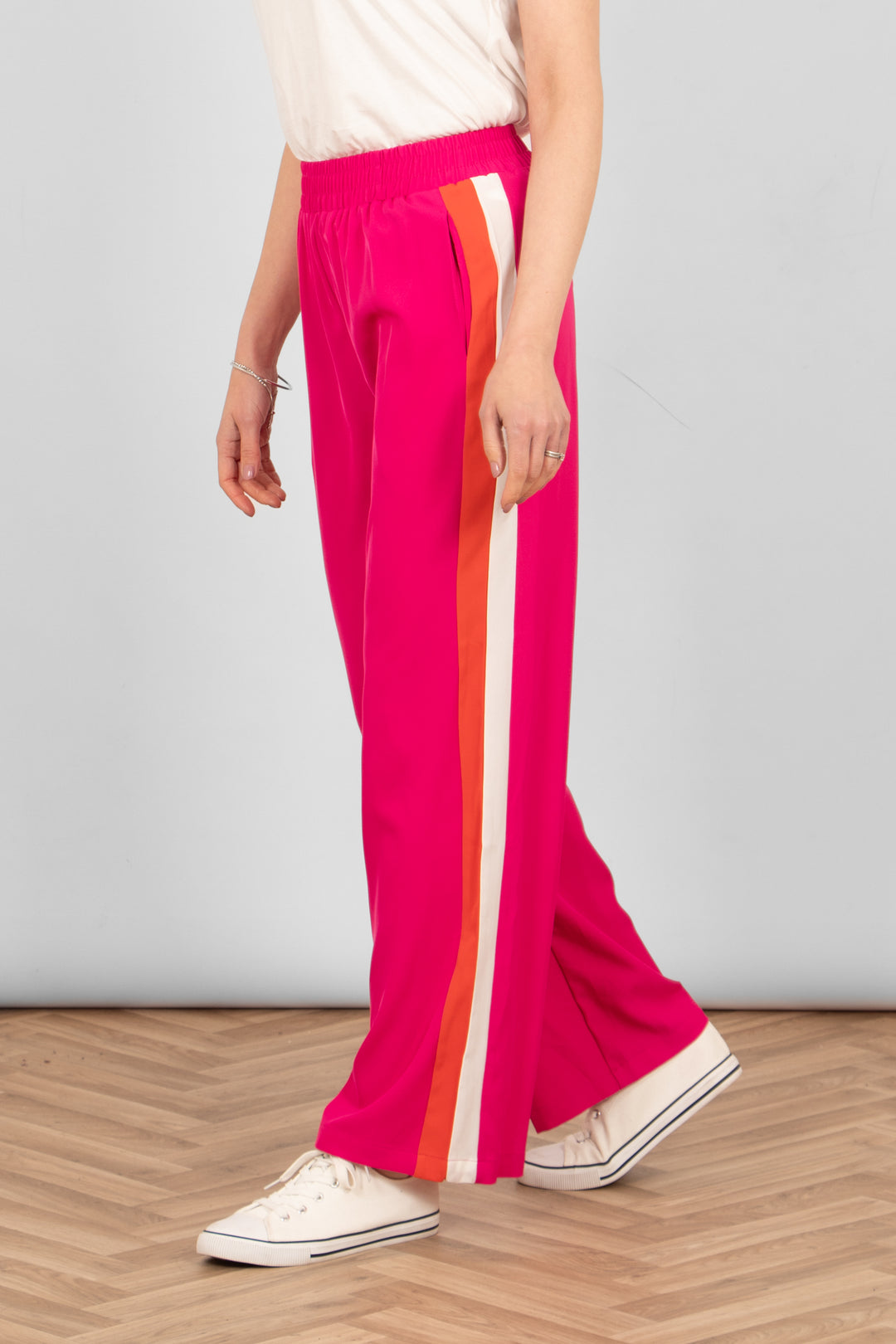 model wearing a pair of hot pink causal trousers with two vertical stripes running down the side of the leg. one stripe is orange the other is white.