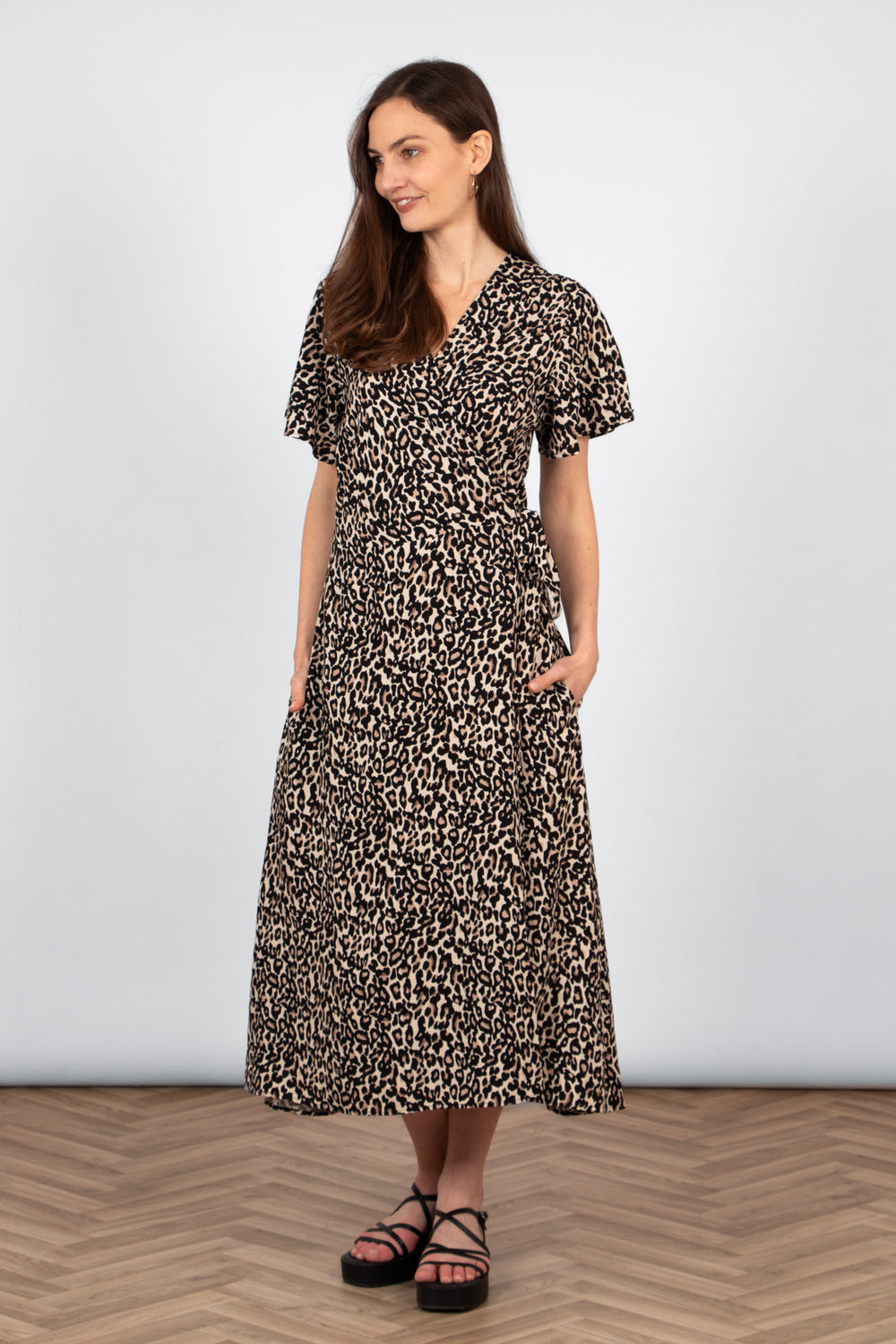 model wearing a neutral leopard print wrap dress. the waist tie is shown at tied in a bow at the side. the dress has a v-neck, short sleeves and side pockets.