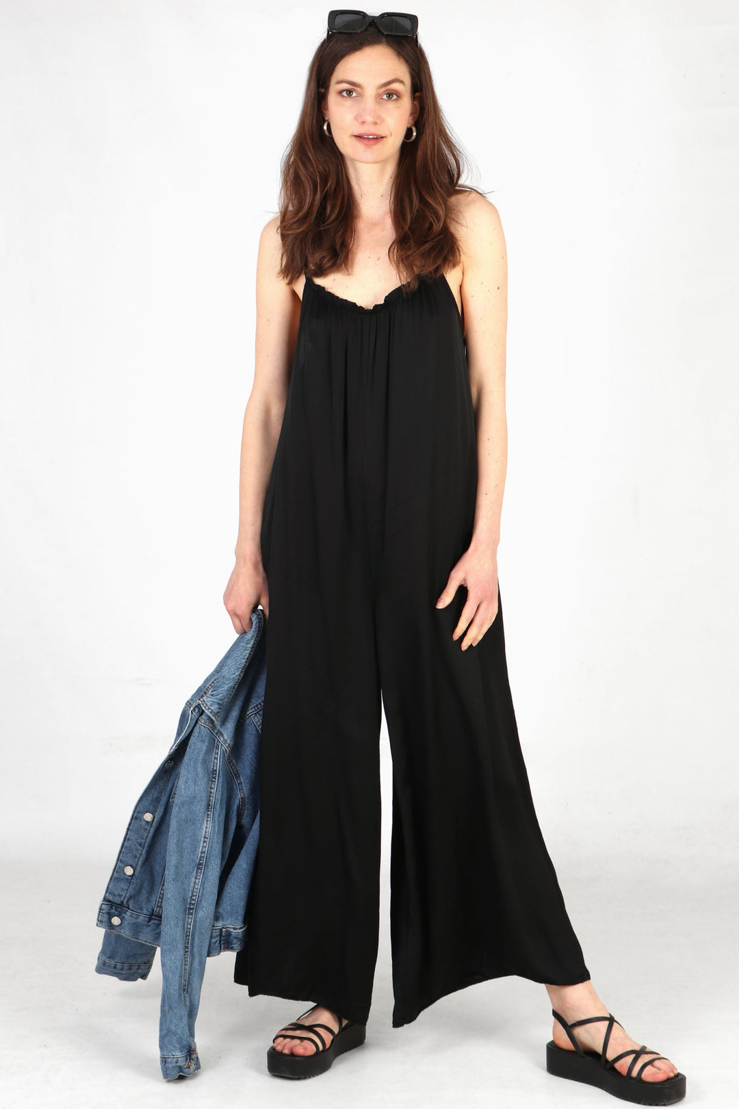 Model wearing black silky texture jumpsuit with denim jacket in hand. Style with black sandals