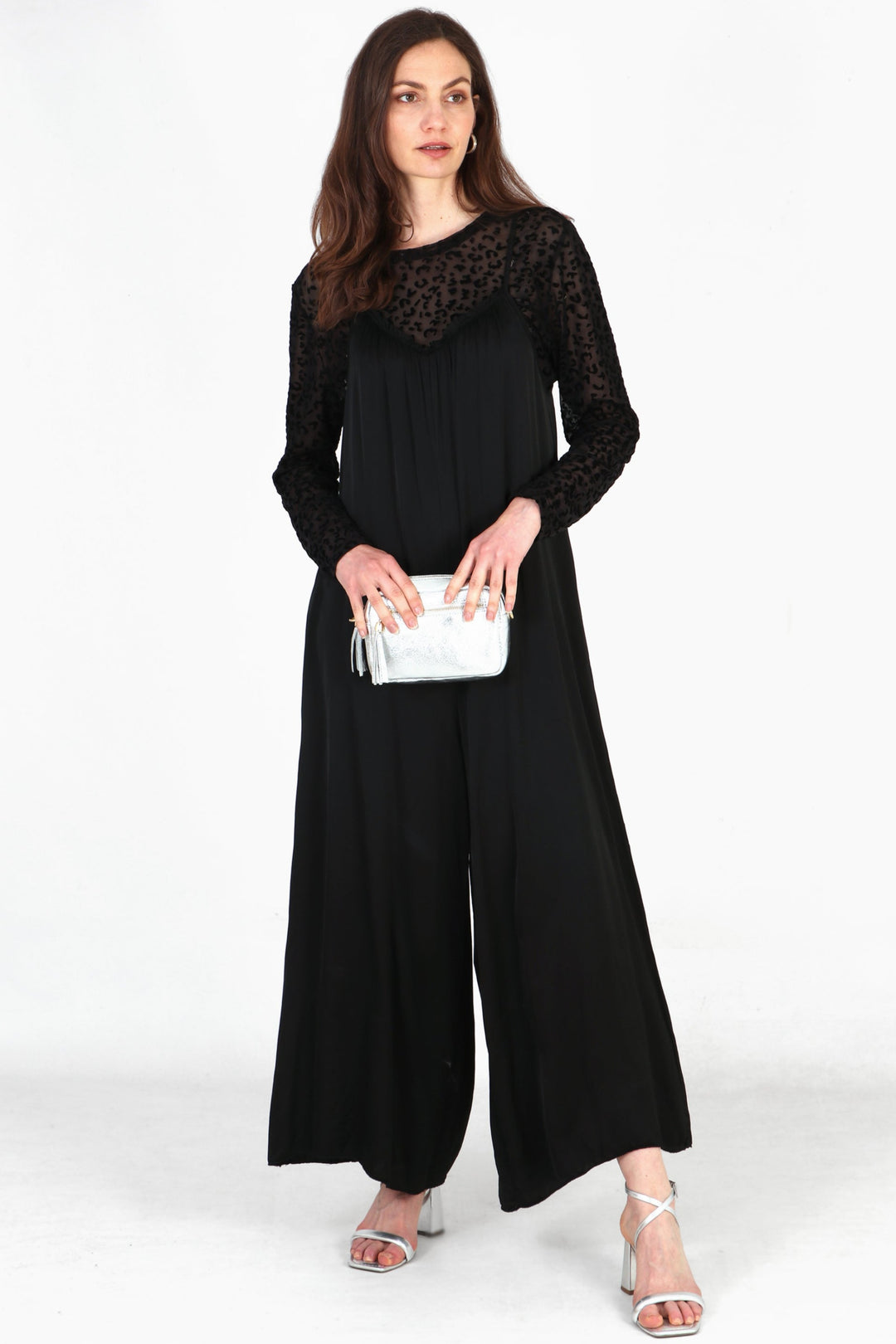 Model wearing black silky strappy wide leg jumpsuit with black sheer leopard print top underneath. Holding silver camer bag and wearing silver heels