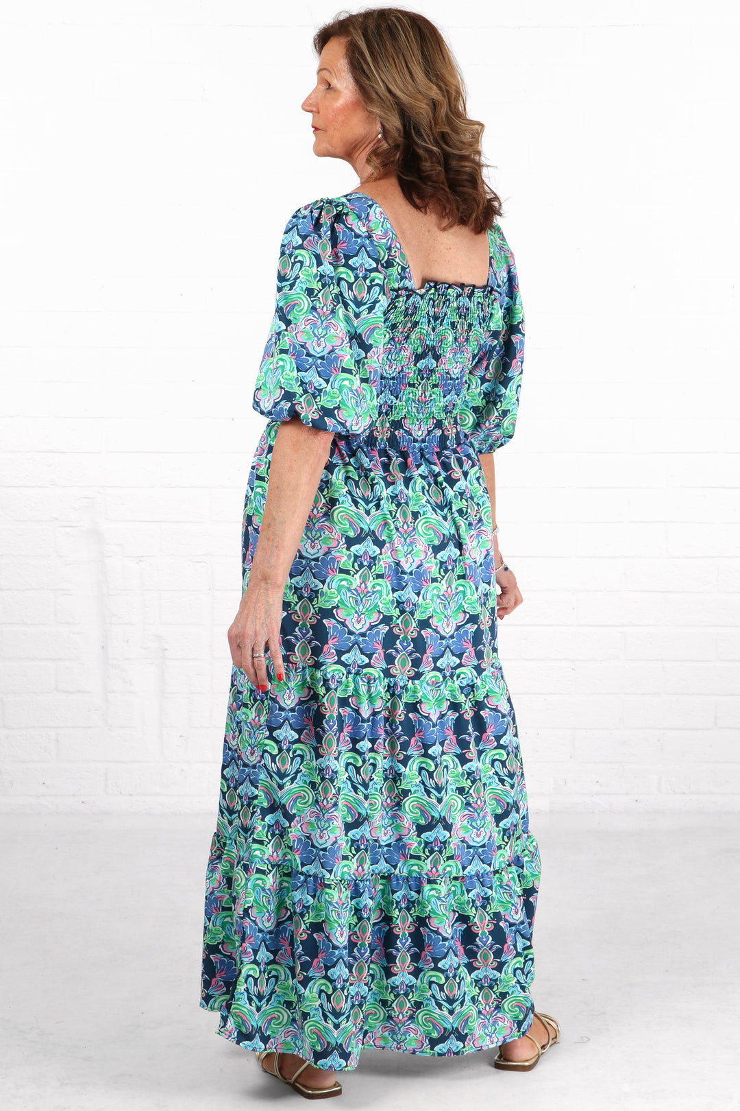 model showing the back of the blue fleur de lis maxi dress, showing a square neck line and shirred bodice