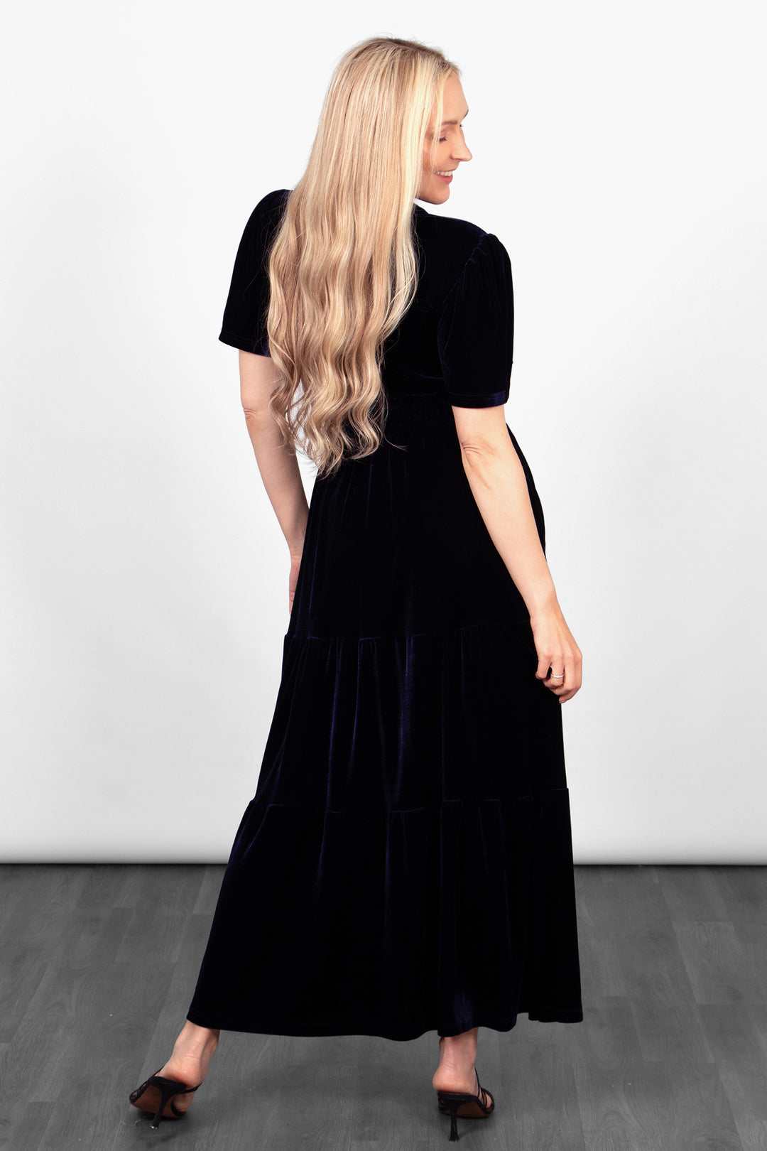 model showing the back of the navy blue velvet dress, showing that the hemline falls at the ankle and this dress is maxi length