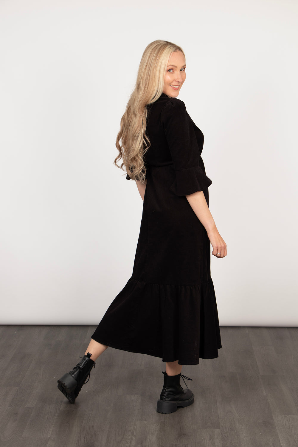 model showing a side view of the black baby cord dress, the fluted sleeves and tiered design are visible. the dress is plain black 