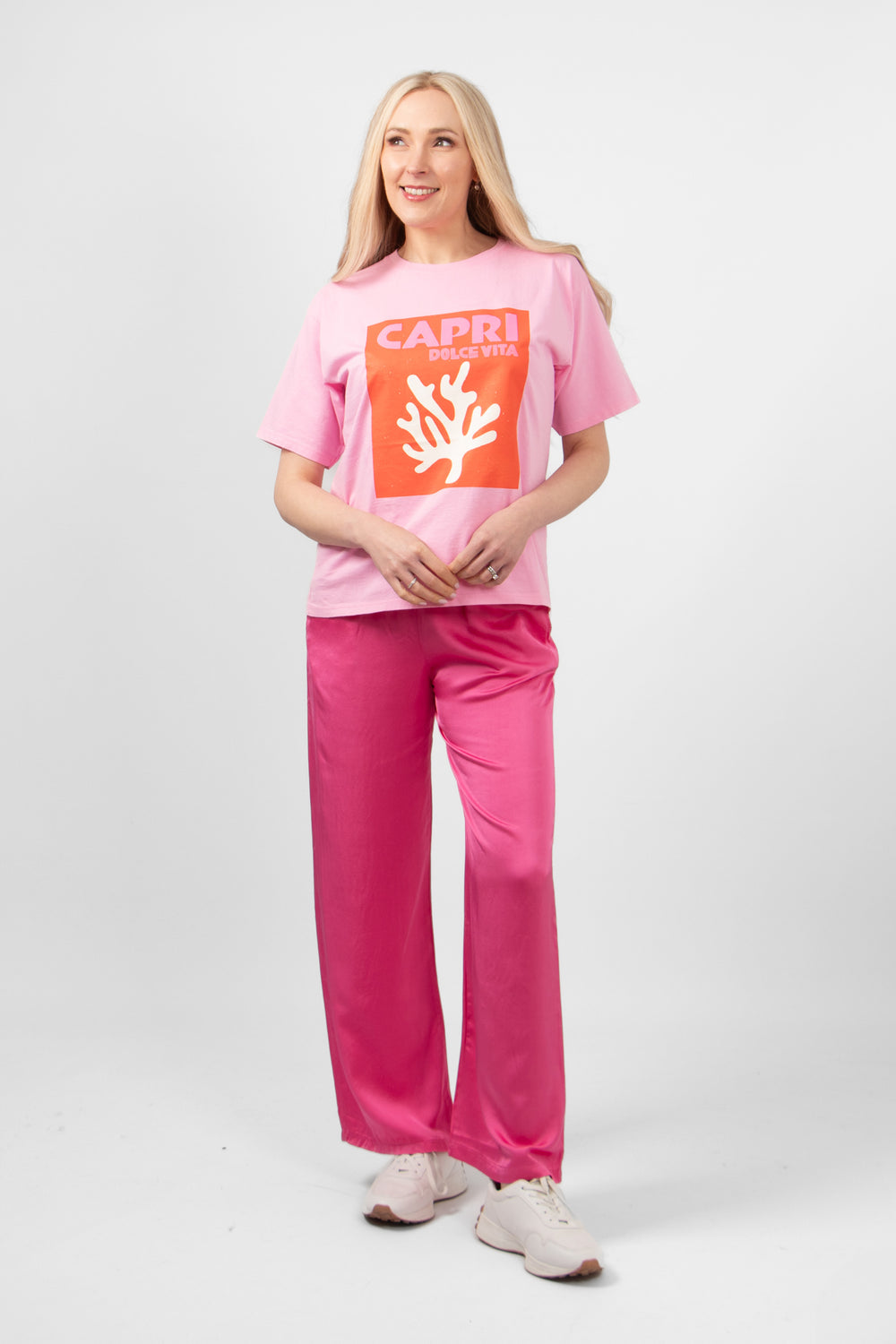 model wearing a pink short sleeves tshirt with a coral motif and the slogan "capri dolce vita" on the front. the tee is styles with pink summer trousers and casual white trainers.