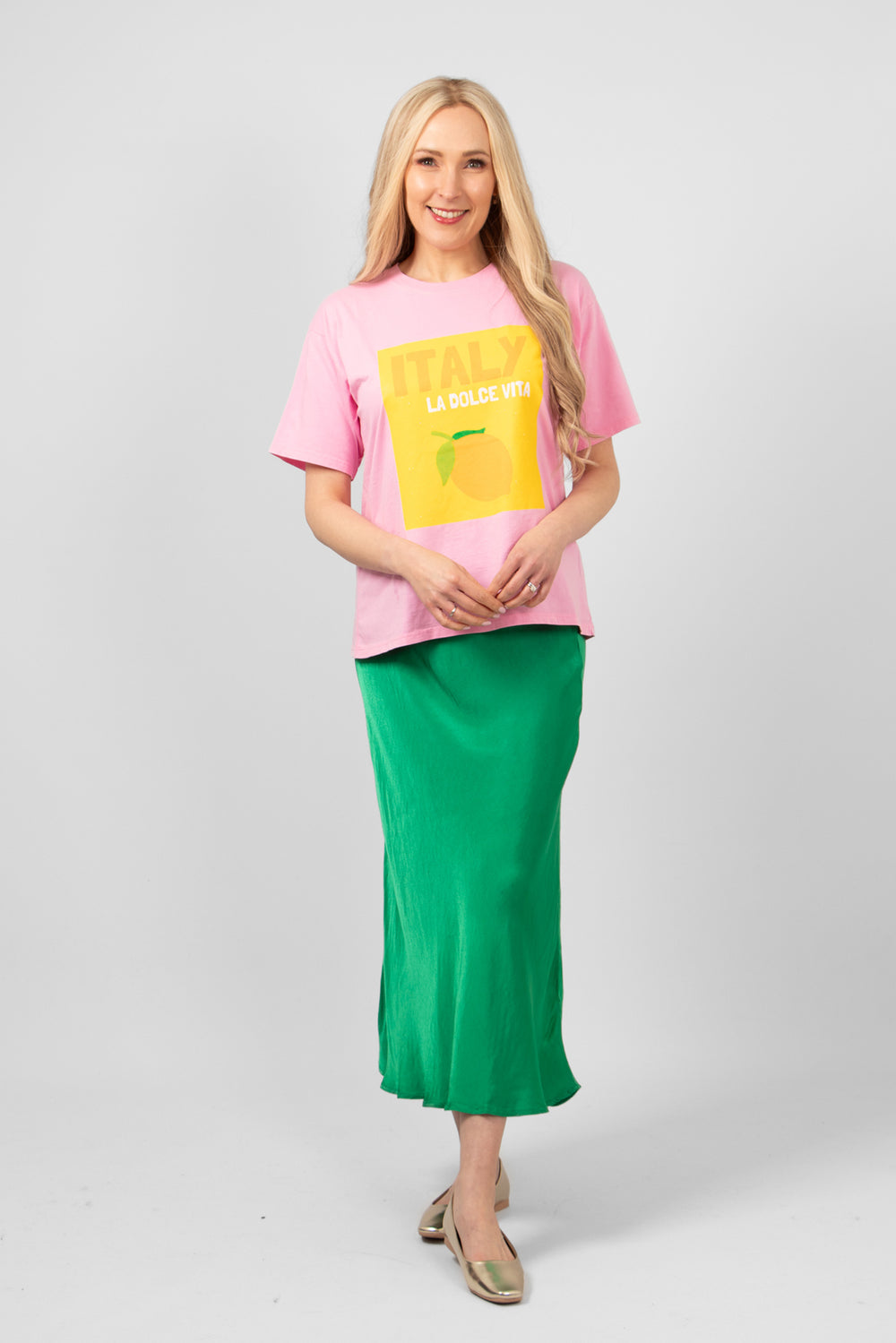 model wearing a pink cotton tshirt with a square yellow graphic logo on the front. the t-shirt is styled with a green midi slip skirt and gold flat pumps.