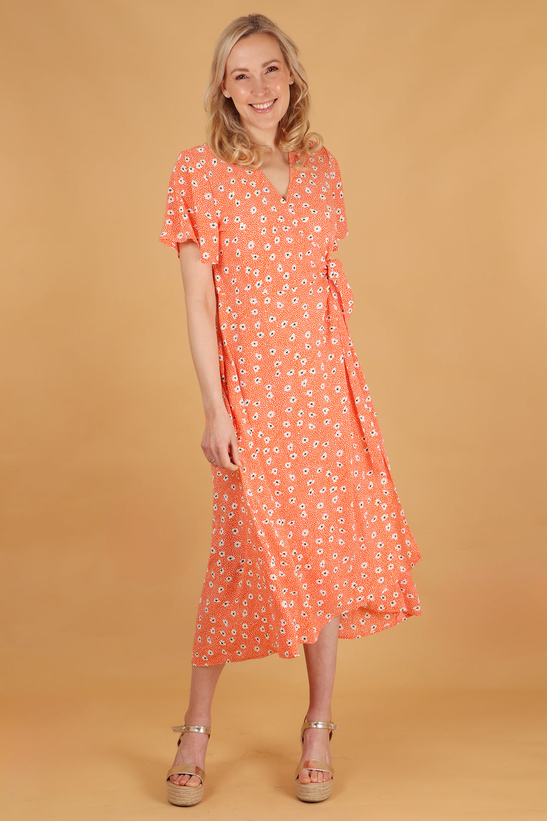 model wearing an orange midi length wrap dress with an all over white daisy floral pattern. the dress has short sleeves, waist tie and vneck